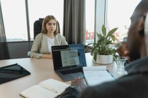 A woman sits across from a professional in an office as they review legal documents on a laptop and notepad.
