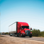 A red semi-truck with a white trailer driving down a highway under a clear blue sky.