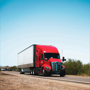 A red semi-truck with a white trailer driving down a highway under a clear blue sky.