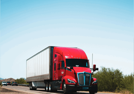 A red semi-truck with a white trailer driving down a highway under a clear blue sky.