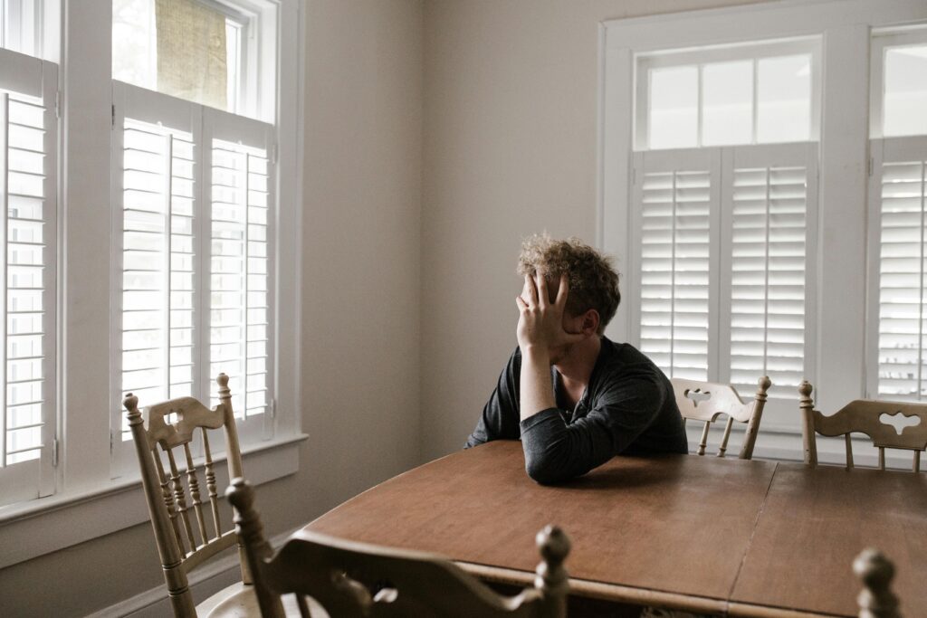 Man sits alone in his home at the kitchen table holding his face in his hands.