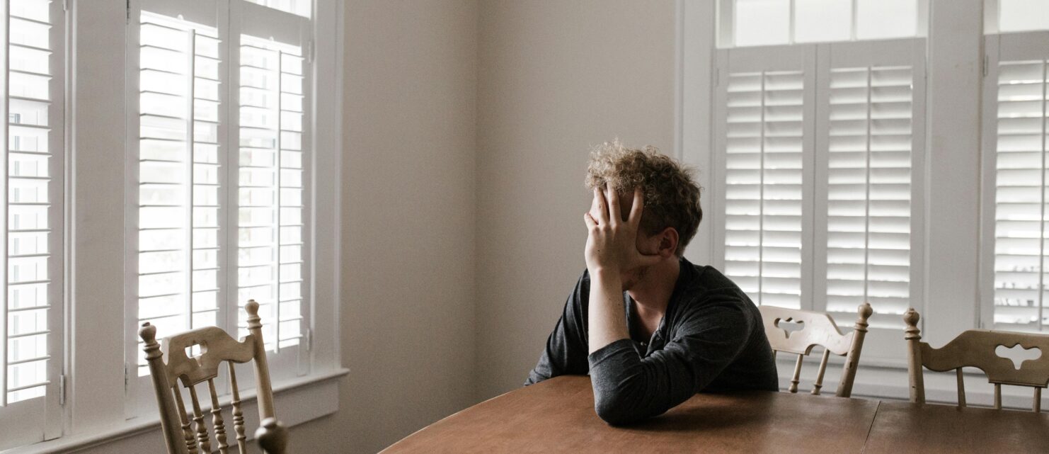 Man sits alone in his home at the kitchen table holding his face in his hands.