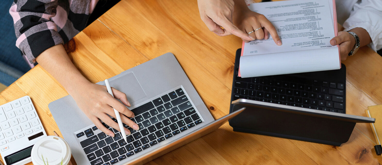 Two people sit at a table looking at paper records and a laptop