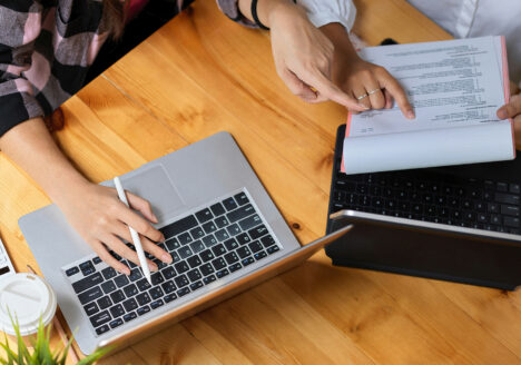 Two people sit at a table looking at paper records and a laptop