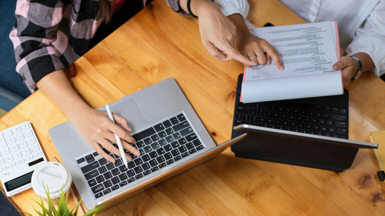 Two people sit at a table looking at paper records and a laptop